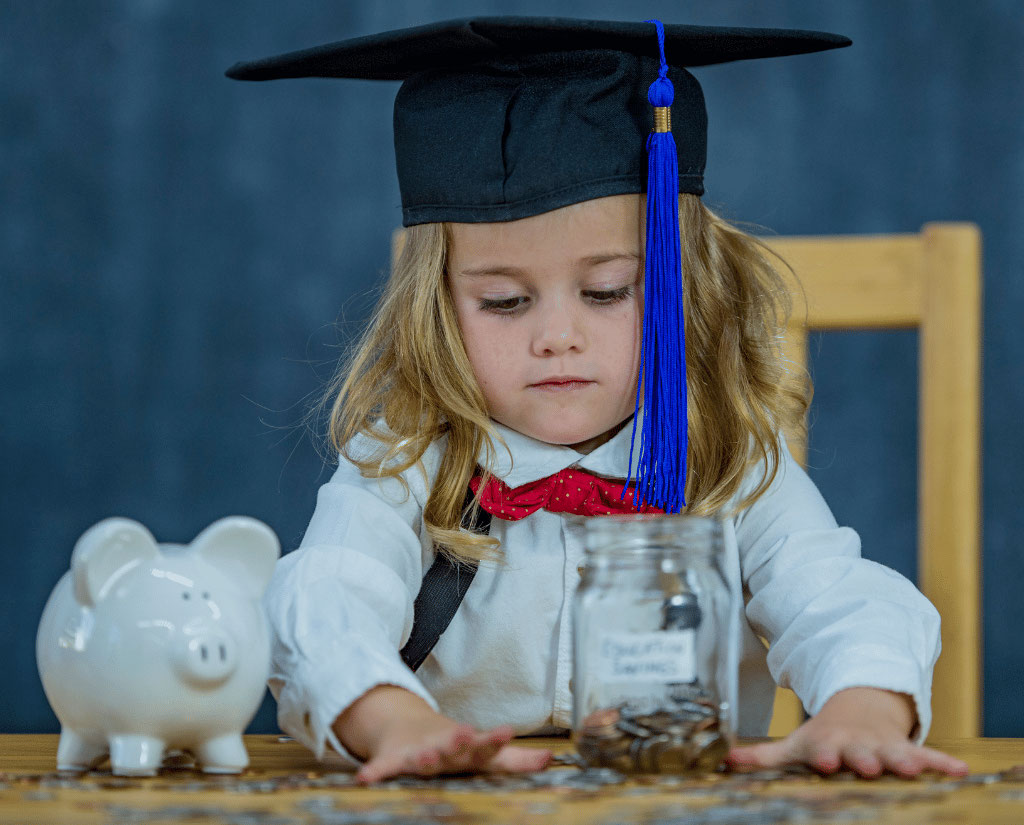 A little girl wears a grad cap and sits at a desk counting the coins from her piggy bank