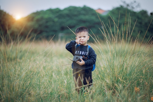 Young boy standing in a field pointing face on while holding a camera