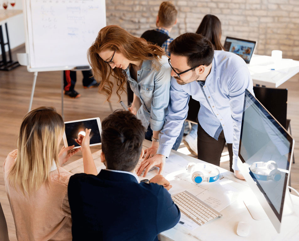 Four people in a shared workspace with two sitting a desk and the others standing up and they are all looking at an ipad with a look of interest, depicting collaboration and a shared idea.