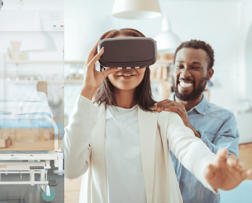 Man smiling while standing behind a woman with his hands on her shoulder and back as she wears Virtual Reality goggles