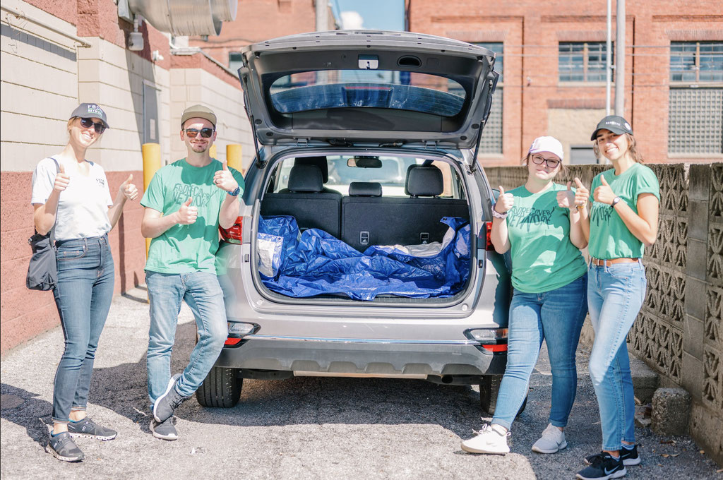 4 people standing behind a car giving a "thumbs-up" . The car's trunk is open